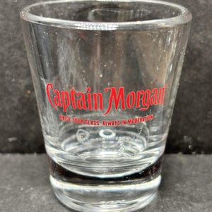 Może przedstawiać: Przezroczysty kieliszek do shotów z czerwonym logo "Captain Morgan" i napisem "Raise your glass. Always in moderation." Kieliszek jest cylindryczny z grubą podstawą, przeznaczony do alkoholi.
