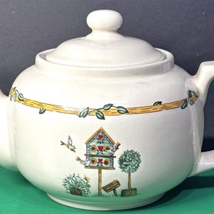 Könnte beinhalten: Eine cremefarbene Keramik-Teekanne mit passendem Deckel, verziert mit einem bemalten Motiv aus einem Vogelhaus, kleinen Vögeln und Grünpflanzen. Ein gelbes horizontales Band mit grünen Blättern umrandet den oberen Teil des Kannenbauchs.
