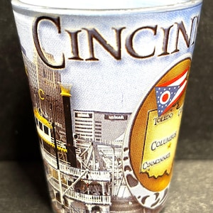 May include: A clear shot glass with a design featuring the city name "CINCIN" in large letters. The glass has a cityscape and a state flag design. The design includes the cities of Toledo, Columbus, and Cincinnati.