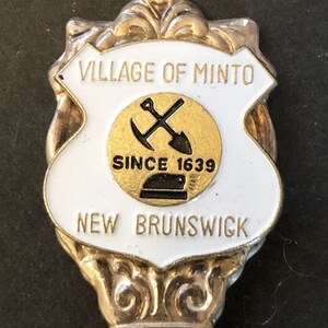 Może przedstawiać: Srebrna łyżeczka pamiątkowa z białą emaliowaną tarczą z napisem "Village of Minto, New Brunswick, Since 1639". Tarcza ma złote koło z przekreślonym kilofem i łopatą.