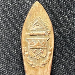 Könnte beinhalten: Ein kupferfarbener Löffel mit spitzem Ende. Der Löffel ist mit einem allsehenden Auge, einer Schriftrolle, einer Bergkette, gekreuzten Spitzhacken und den Worten "In Unitate Fortis Sine." graviert.