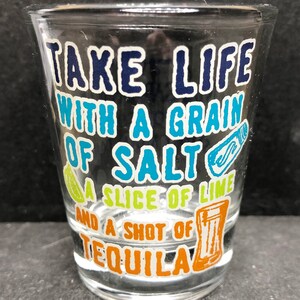 Pode incluir: Um copo de shot de vidro transparente com o texto "TAKE LIFE WITH A GRAIN OF SALT A SLICE OF LIME AND A SHOT OF TEQUILA" impresso nele em azul, verde, amarelo e laranja.