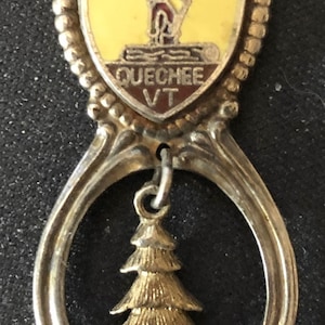 May include: A silver spoon with a gold-colored charm. The charm is a shield with the words "Timber Village" and "Queechee VT". A small gold-colored pine tree hangs from the charm.