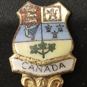 May include: A gold-colored souvenir spoon with a white enamel design. The design features the Canadian coat of arms with a red and white shield, a blue and white harp, and three green maple leaves. The spoon has a white enamel banner with the word "CANADA" in black lettering.