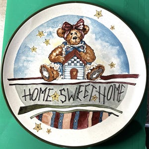 Puede incluir: Un plato decorativo con un diseño caprichoso. El plato presenta un oso de peluche marrón con un peto azul y un lazo rojo, sosteniendo una pequeña casa. Las palabras "HOME SWEET HOME" están escritas en el plato.