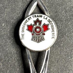 Puede incluir: Marcapáginas plateado con un emblema circular. El emblema presenta un tren rojo y un diseño de hoja de arce, con el texto "THE DISCOVERY TRAIN - LA DÉCOUVERTE". El marcapáginas tiene una forma curva y alargada.