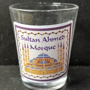 Puede incluir: Un vaso de chupito transparente con una etiqueta que representa la Mezquita del Sultán Ahmed. La etiqueta tiene un borde morado con un diseño floral. La mezquita está ilustrada en azul, dorado y marrón.