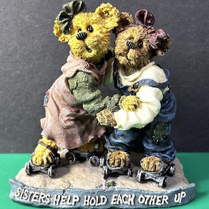 May include: A figurine of two teddy bears embracing, with the text "Sisters Help Hold Each Other Up" on the base. One bear wears a pink dress and the other wears blue overalls. The bears are on a stone-like base.