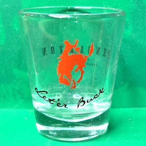 May include: Clear shot glass with a red bucking bronco graphic and the words "Let'er Buck" in black script. The word "PENTLETON" is printed in reverse. The glass is on a green surface.