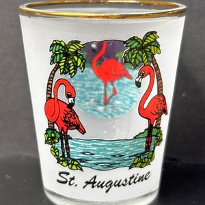 Peut inclure: Un verre à shot en verre dépoli avec un bord doré, orné d'un motif représentant trois flamants roses, des palmiers verts et une étendue d'eau bleue. Le texte St. Augustine est imprimé en écriture noire à la base du verre.