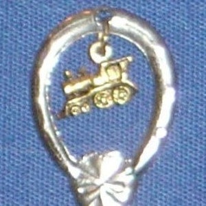 Puede incluir: Cuchara de plata con un dije de tren de color dorado colgando de ella. La cuchara tiene un mango en forma de lazo.