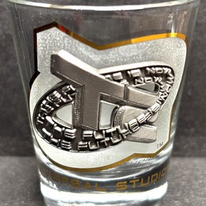 May include: Clear shot glass with a silver and gold emblem featuring the text "The Future is Now" and "Universal Studios". The glass has a cylindrical shape and a thick base. The emblem is metallic and has a raised design.