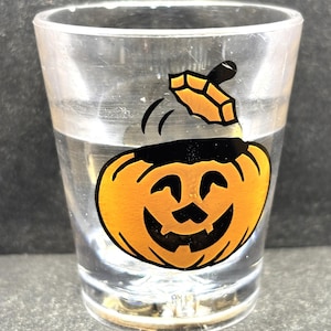 May include: Clear shot glass with a smiling jack-o'-lantern design in orange and black. The glass is filled with a clear liquid. The pumpkin graphic has a black stem and a smiling face. Perfect for Halloween.