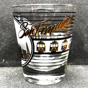 May include: A clear shot glass with a San Francisco design. The glass features the city's name in gold script, cable cars, and a skyline silhouette. Black and white horizontal lines and a black border complete the design.