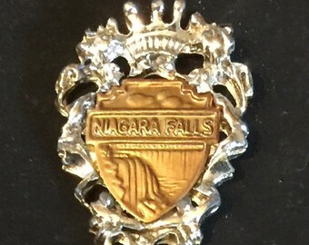 Könnte beinhalten: Ein silberner Löffel mit einem goldfarbenen Anhänger, der ein Wappen mit den Worten "NIAGARA FALLS" und einer Darstellung der Fälle zeigt. Der Anhänger ist von einem dekorativen silbernen Rahmen umgeben.