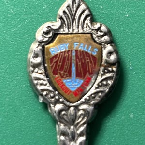 May include: A silver-toned souvenir spoon with an ornate handle and a shield-shaped emblem. The emblem features the words "Ruby Falls Lookout Mtn" in red and blue, with a waterfall illustration.