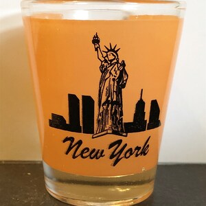 Könnte beinhalten: Orangenes Glas-Schnapsglas mit einer schwarzen Silhouette der Freiheitsstatue und der Skyline von New York. Der Text "New York" ist unter dem Bild gedruckt.