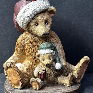 Puede incluir: Figura de oso marrón con un oso más grande sentado detrás de un oso más pequeño. Ambos osos llevan gorros de Papá Noel con ribetes blancos y bayas de acebo. El oso más pequeño sostiene una caja de regalo roja. La figura está sobre una base marrón con aspecto de madera.