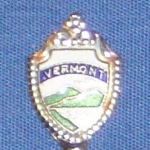 May include: A gold-colored souvenir spoon with a blue and white enamel design. The spoon features a shield-shaped design with the word "VERMONT" and a depiction of mountains and a river.