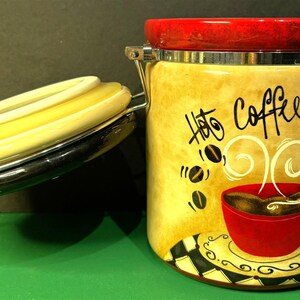 May include: A ceramic canister with a hinged lid and a rubber seal. The canister features a tan background with a graphic of a red coffee cup, steam, and coffee beans. The text "Hot Coffee" is printed in black script on the front of the canister.