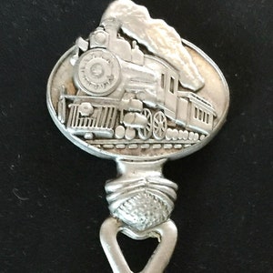 Puede incluir: Cuchara de plata con un diseño detallado de un tren de vapor con humo saliendo de la chimenea. El tren está en una vía y tiene varios vagones. El mango de la cuchara tiene forma de nudo.