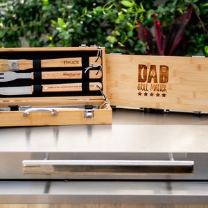 May include: A wooden box with a set of grilling tools inside. The box is engraved with the words "Dad Grill Master" and five stars. The tools include a spatula, tongs, a fork, and a brush.