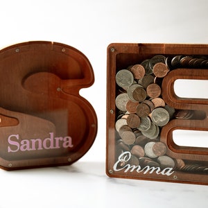 May include: Two wooden piggy banks shaped like the letters "S" and "E". The "S" piggy bank has the name "Sandra" written on it in pink letters. The "E" piggy bank has the name "Emma" written on it in white letters. Both piggy banks have coins inside.