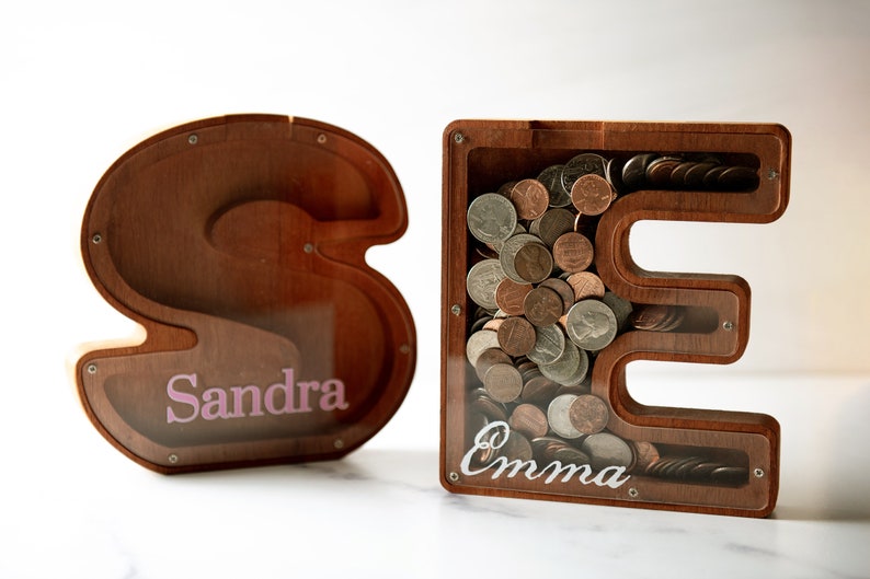 May include: Two wooden piggy banks shaped like the letters "S" and "E". The "S" piggy bank has the name "Sandra" written on it in pink letters. The "E" piggy bank has the name "Emma" written on it in white letters. Both piggy banks have coins inside.