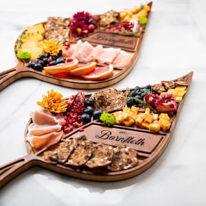 Könnte beinhalten: Zwei hölzerne, blattartige Servierbretter mit Griffen. Die Bretter sind in Abschnitte unterteilt und mit verschiedenen Lebensmitteln gefüllt, darunter Käse, Cracker, Obst und Fleisch. Die Bretter sind mit dem Text "Barnfleth" graviert.