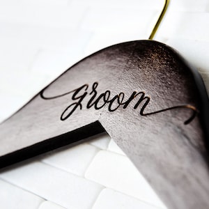 May include: A dark brown wooden hanger with a gold hook and the word "groom" engraved in a cursive font.