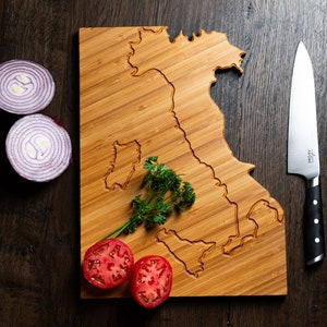 Könnte beinhalten: Ein Holzbrett in Form der Umrisse Italiens, mit einem Messer, roten Zwiebelhälften, Tomatenhälften und Petersilie darauf.