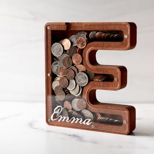 May include: A wooden piggy bank shaped like the letter "E" with a clear acrylic front. The bank is filled with coins and has the name "Emma" inscribed on the front.