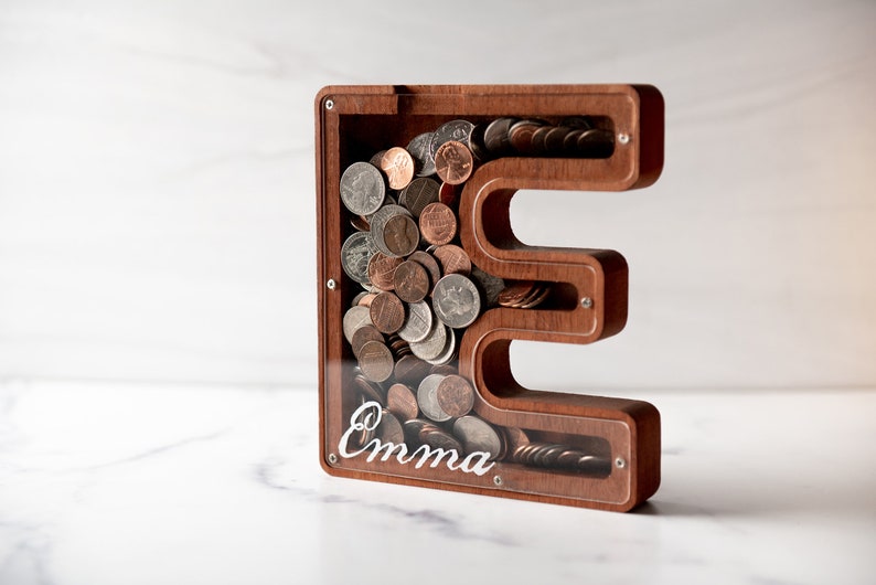 May include: A wooden piggy bank shaped like the letter "E" with a clear acrylic front. The bank is filled with coins and has the name "Emma" inscribed on the front.