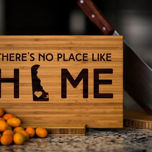 May include: A wooden cutting board with the text "There's no place like home" and an outline of the state of Delaware with a heart in the center. The board is sitting on a counter with a pile of small orange fruits in front of it.