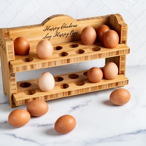 Könnte beinhalten: Ein zweistöckiger Bambus-Eierhalter mit dem Text "Happy chickens lay Happy Eggs" eingraviert auf der Oberseite. Der Halter hat 18 Fächer für Eier und ist mit braunen Eiern gefüllt.