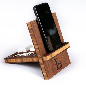 Op de afbeelding: Een houten telefoonstandaard met een oplaadstation en een gepersonaliseerde inscriptie met de tekst "LENNOX L". De standaard is gemaakt van donkerbruin hout met een lichtbruine accent.