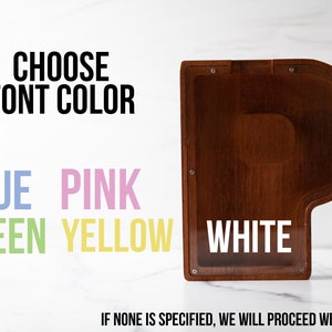May include: A wooden piano-shaped box with a clear plastic window. The text "CHOOSE FONT COLOR" is above a list of color options: blue, pink, green, yellow, and white. The text "IF NONE IS SPECIFIED, WE WILL PROCEED WITH WHITE" is below the color options.