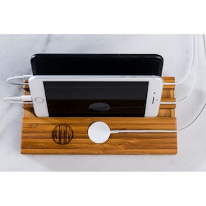 May include: A bamboo charging station with two slots for phones and a space for a watch. The station has a white charging cable and a white wireless charging pad. The station is engraved with the letters "THE" in a circle.