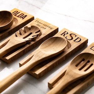 May include: Four wooden spoon rests with personalized names and a saying "I love stirring the pot". Each rest holds a wooden spoon or spatula.