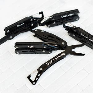 May include: Five black multi-tool pocket knives with engraved text. The knives have a variety of tools, including a blade, pliers, and a bottle opener. The text on the knives reads "Groom", "Groomsman", "Best Man", "ADS", and "Charles".