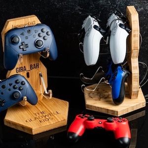 May include: A wooden stand with the text "The Gladwell Kids" engraved on it. The stand holds four video game controllers, two blue and two white. The stand is on a black surface.