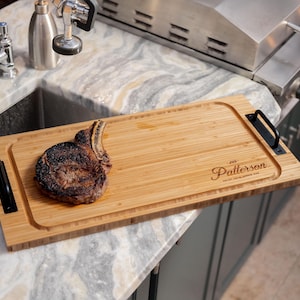 Puede incluir: Un filete de costilla a la parrilla sobre una tabla de cortar rectangular de bambú con asas negras. La tabla tiene el texto grabado "Patterson" y está colocada sobre una encimera de mármol. El filete está cocinado a punto.