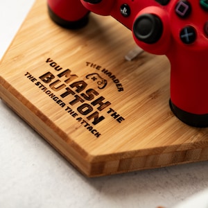 Op de afbeelding: Een rode en zwarte gamecontroller rust op een bamboe houten standaard. De standaard is gegraveerd met de tekst "YOU MASH THE BUTTON THE HARDER THE ATTACK THE STRONGER THE ATTACK". De controller heeft een levendige rode kleur.