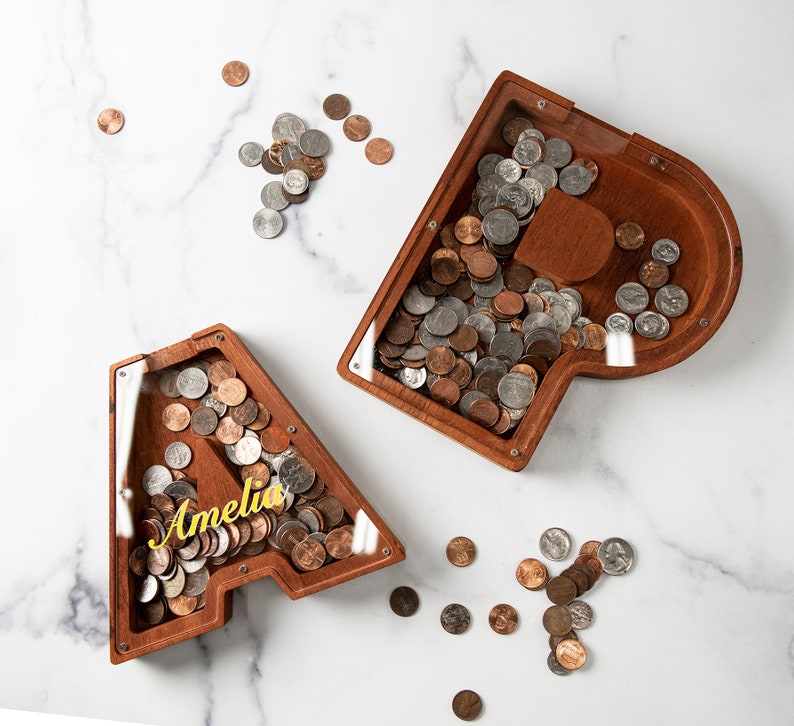 May include: Two wooden piggy banks shaped like the letters A and P, with a glass front to see the coins inside. The A piggy bank has the name Amelia written on it. Both piggy banks are filled with coins.