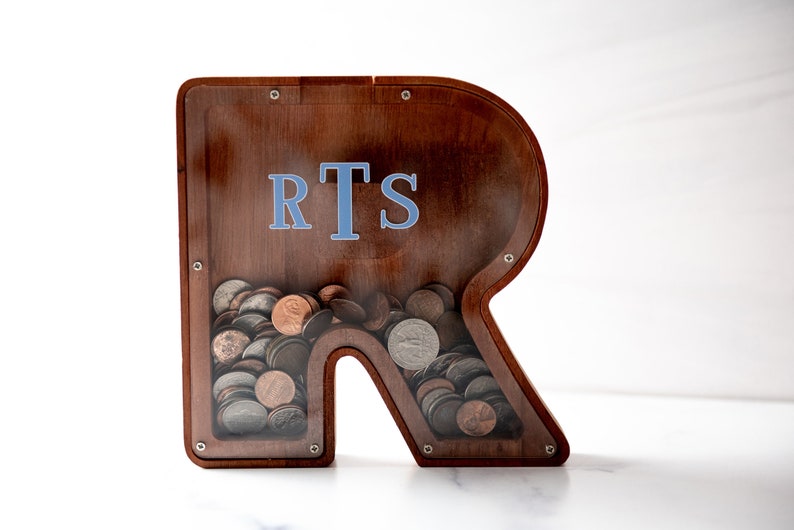 May include: A wooden piggy bank shaped like the letter R with a clear plastic front. The piggy bank has the initials "RTS" written on the front in blue letters. The piggy bank is filled with coins.