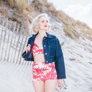 Peut inclure: Une femme portant une veste en jean bleu sur un haut de bikini floral rouge et blanc et un bas de bikini taille haute assorti. Elle se tient sur une plage de sable avec une clôture en bois derrière elle.