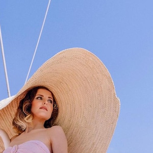 Op de afbeelding: Een vrouw draagt een roze gingham bikini en een grote, breedgerande strohoed. De hoed is beige en biedt schaduw. De achtergrond is een helderblauwe lucht en een witte reling.