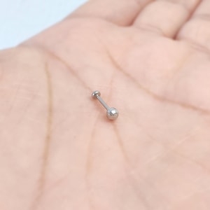 May include: A small, silver-colored barbell earring with a flat disc on one end and a faceted ball on the other. The earring is shown against a light pink background.