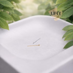 Puede incluir: Dos piercings de nariz, uno plateado y otro dorado, se muestran sobre una superficie blanca. El piercing dorado tiene un pequeño extremo de bola, mientras que el piercing plateado tiene un extremo de bola similar. El fondo presenta follaje verde y el texto "LF4Y CRAFTED BODY JEWELLERY".