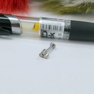 Könnte beinhalten: Ein silberfarbenes Lippenpiercing mit drei klaren, runden Edelsteinen. Das Piercing hat einen flachen Rücken und einen geraden Stab. Ein schwarzer Stift mit transparentem Schaft und Barcode befindet sich im Hintergrund.